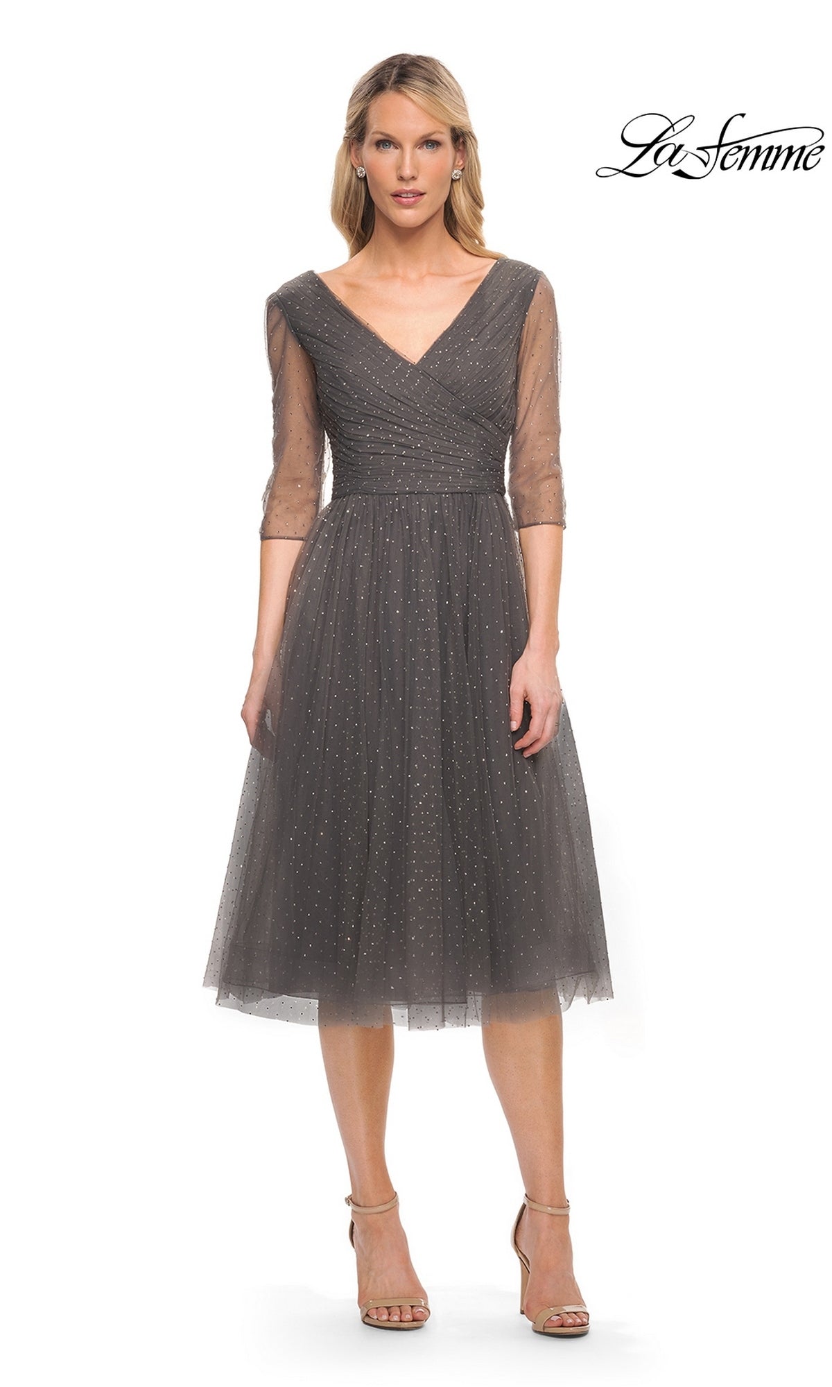 Woman in Knee Length Wedding Guest Dress: La Femme 29229 in gunmetal with sheer sleeves and sparkling v-neckline, white background, “La Femme” logo top right.